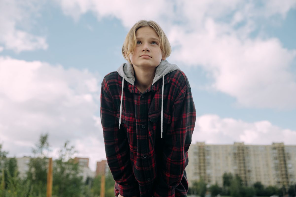 A blond teenage boy with a modern casual haircut looks into the distance outdoors.