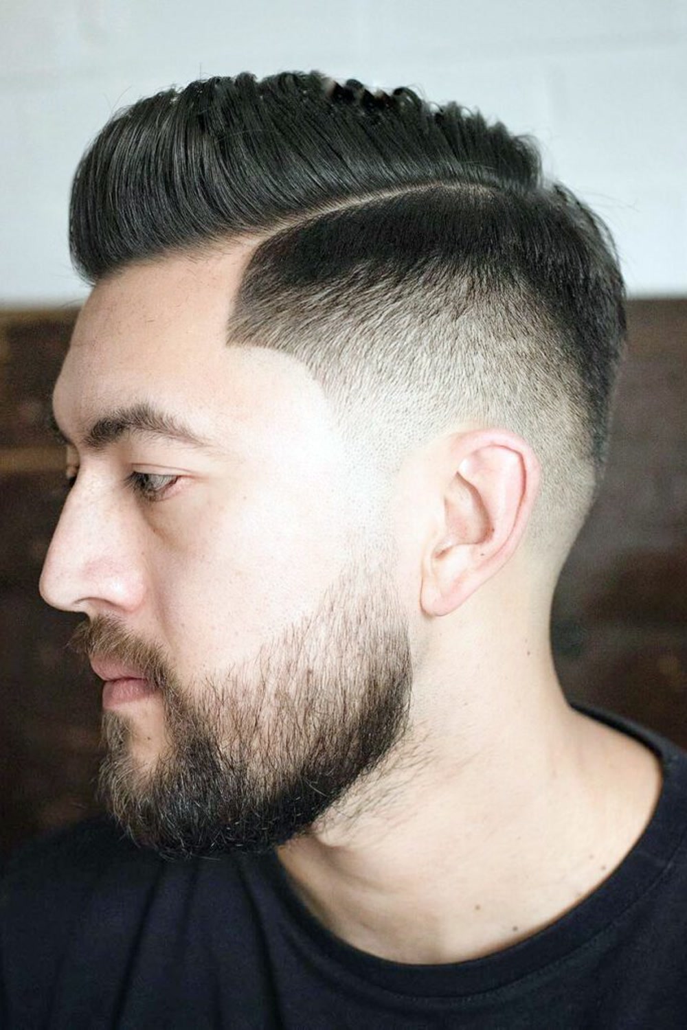 Young man with a burst fade and short pompadour — crisp faded curve around the ear, full beard